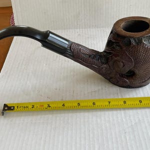 May include: A brown wooden pipe with a black stem and intricate carvings. The pipe is about 7 inches long.