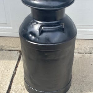 May include: A tall, cylindrical metal milk can finished in a solid matte black paint. The container features a wide, circular lid at the top, a narrow neck, and a slightly wider body with a small handle attached to the shoulder area of the vessel.