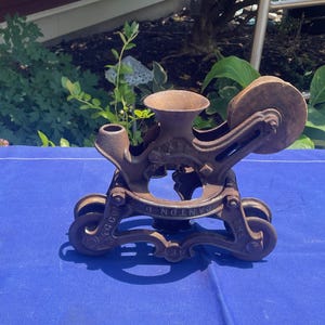 May include: A vintage cast iron meat grinder with a brown, weathered finish. The base features embossed text reading THE NEY MFG CO CANTON O. The device includes a central funnel, a side-mounted wooden wheel, and a sturdy frame with four small wheels.