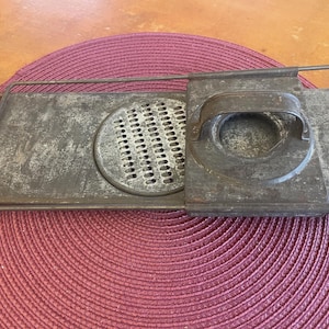 May include: An antique metal grater with a rectangular frame and a circular grating surface. The grater has a handle and a sliding component. The metal has a worn, aged appearance, suggesting it is vintage.