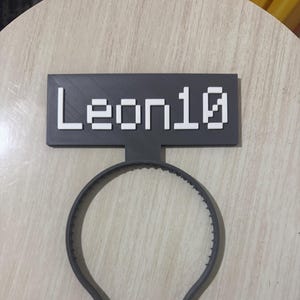 May include: A gray plastic headband with a gray rectangular sign attached to the top. The sign has the word "Leon10" printed in white.