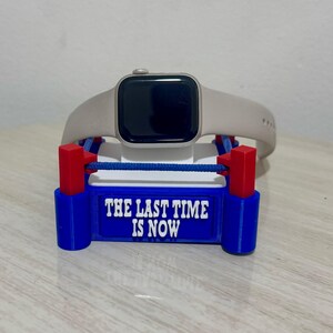 Pode incluir: Um Apple Watch prateado com uma banda branca está sentado em um suporte de impressão 3D azul e vermelho. O suporte tem a forma de um ringue de boxe e tem as palavras "The Last Time Is Now" impressas na frente.