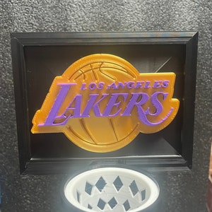 Puede incluir: Un logotipo de los Los Angeles Lakers en oro y morado con un diseño de baloncesto. El logotipo está enmarcado en un marco negro.