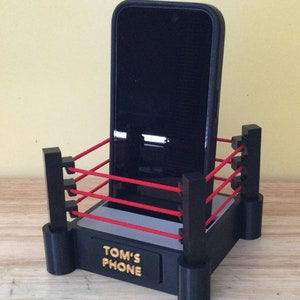 Könnte beinhalten: Ein schwarzer Telefonständer in Form eines Boxrings mit roten Seilen. Der Ständer hat eine schwarze Basis mit den Worten "TOM'S PHONE" in goldenen Buchstaben.