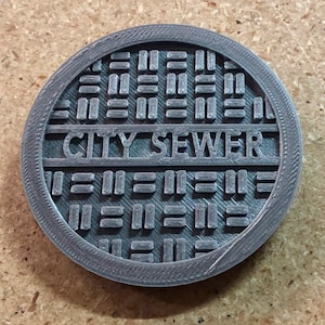 May include: A gray, circular, 3D printed manhole cover with the words "CITY SEWER" embossed on the surface.