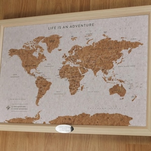 May include: A framed world map made of cork, with a light wood frame. The map shows continents in brown against a light background. The text "LIFE IS AN ADVENTURE" is printed at the top. A small metal tag is at the bottom.