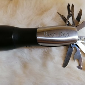 May include: A black flashlight with a silver multi-tool attached. The multi-tool has a variety of blades and tools, including a bottle opener, a saw, and a screwdriver. The flashlight has a silver band around it that says "Best Dad".