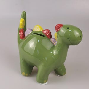 May include: A green ceramic dinosaur-shaped piggy bank with colorful accents. The dinosaur has a smiling face, a slot for coins, and colorful spikes along its back and tail. The piggy bank is approximately 8 inches tall.