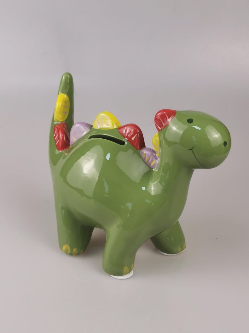 May include: A green ceramic dinosaur-shaped piggy bank with colorful accents. The dinosaur has a smiling face, a slot for coins, and colorful spikes along its back and tail. The piggy bank is approximately 8 inches tall.