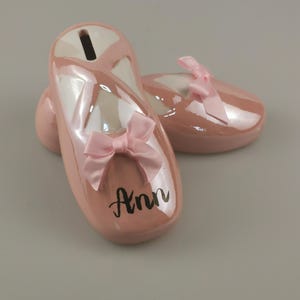Könnte beinhalten: Ein Paar rosa Keramik-Sparschuhe in Form von Ballettschuhen. Jeder Schuh hat eine rosa Satinschleife und den Namen "Ann" in schwarzer Schrift. Die Sparschuhe haben eine glänzende Oberfläche und einen Schlitz für Münzen.