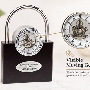 May include: A black rectangular clock shaped like a padlock with a silver shackle. The clock face features Roman numerals and visible moving gears. An oval silver plaque is engraved with the text: Love you to the moon and back LYC Gifts Engraved.
