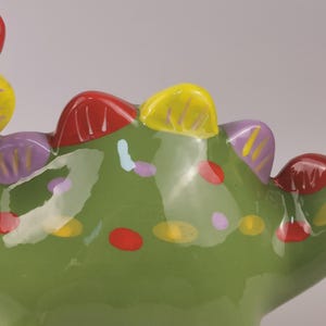 May include: A close-up of a green ceramic decorative object with colorful accents. The object has a textured surface with red, yellow, and purple details. The design includes painted dots and shapes.