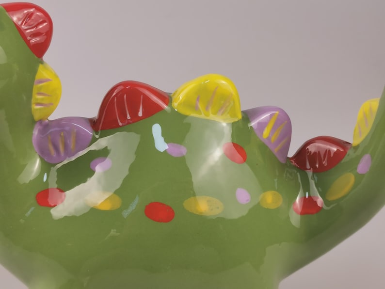 May include: A close-up of a green ceramic decorative object with colorful accents. The object has a textured surface with red, yellow, and purple details. The design includes painted dots and shapes.