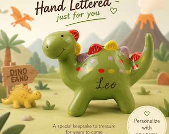 May include: A green ceramic dinosaur piggy bank featuring colorful spots and spikes along its back. The name Leo is written in black script on its side. The text Hand Lettered just for you and Personalize with any name appear in the image.