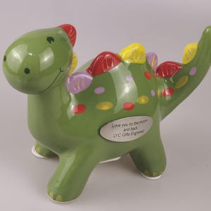 May include: A green ceramic dinosaur figurine with red, yellow, and purple accents. The dinosaur has a smiling face and a small silver plaque that reads "Love you to the moon and back, LYC Gifts Engraved."