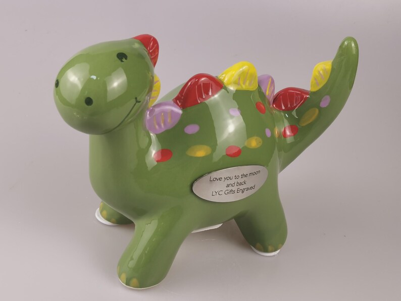 May include: A green ceramic dinosaur figurine with red, yellow, and purple accents. The dinosaur has a smiling face and a small silver plaque that reads "Love you to the moon and back, LYC Gifts Engraved."