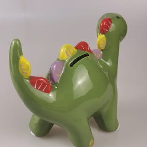 May include: A green ceramic dinosaur-shaped piggy bank with colorful accents. The dinosaur has a long neck and tail, with a coin slot on its back. The accents include red, yellow, and purple details.