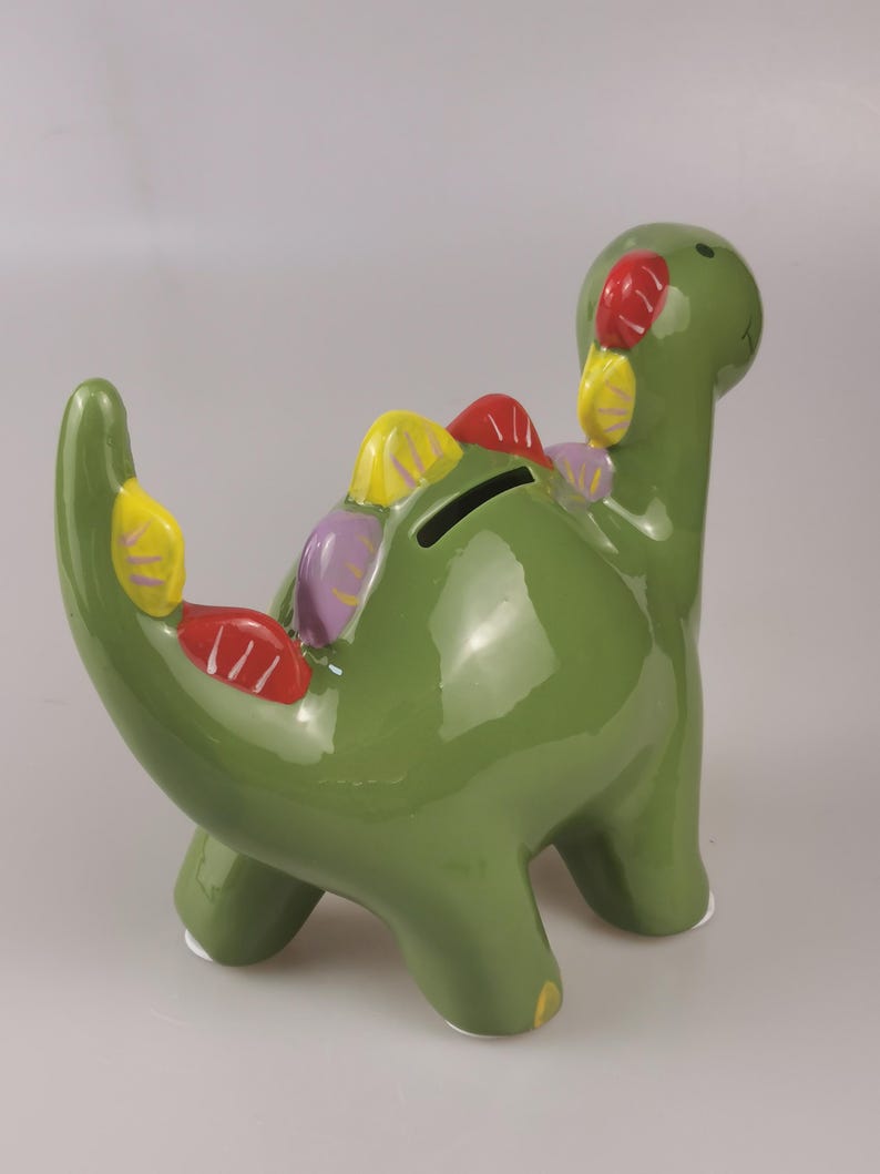 May include: A green ceramic dinosaur-shaped piggy bank with colorful accents. The dinosaur has a long neck and tail, with a coin slot on its back. The accents include red, yellow, and purple details.