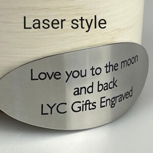 May include: A cream-colored cylindrical object with a silver oval tag. The tag is engraved with the words "Love you to the moon and back" and "LYC Gifts Engraved". The text "Laser style" is above the tag.