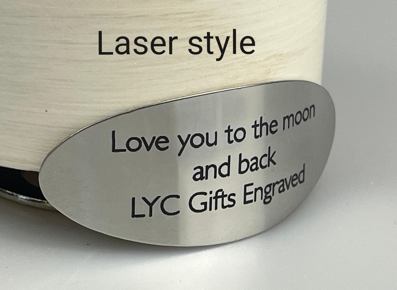 May include: A cream-colored cylindrical object with a silver oval tag. The tag is engraved with the words "Love you to the moon and back" and "LYC Gifts Engraved". The text "Laser style" is above the tag.