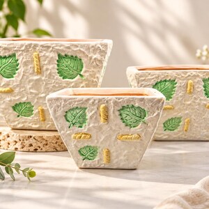 Peut inclure: Trois jardinières carrées blanc cassé avec une surface texturée et froissée. Chaque pot présente des motifs de feuilles vertes en relief et de petits accents rectangulaires jaunes en relief disposés en motif répétitif.