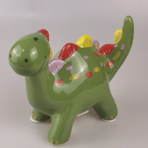May include: A green ceramic dinosaur figurine with a glossy finish. The dinosaur has red, yellow, and purple accents along its back and tail. It has a smiling face and white feet. A decorative item.