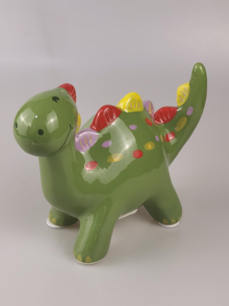 May include: A green ceramic dinosaur figurine with a glossy finish. The dinosaur has red, yellow, and purple accents along its back and tail. It has a smiling face and white feet. A decorative item.