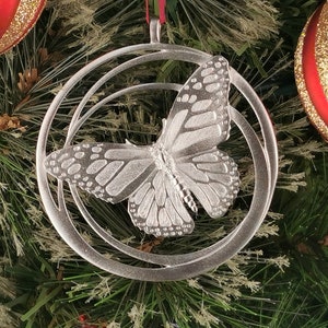 May include: A silver butterfly Christmas ornament with intricate wing detailing, set within a circular frame. The decoration hangs from a red ribbon against a Christmas tree with red and gold baubles.