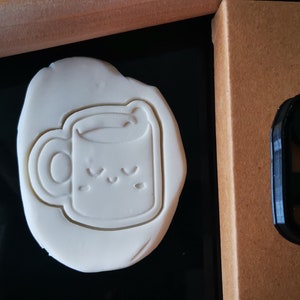 Pode incluir: Um cortador de biscoitos de plástico preto em forma de caneca com um rosto sorridente. O cortador está sobre uma superfície marrom. Uma peça branca de massa de modelar foi pressionada com o cortador, mostrando a impressão da caneca.