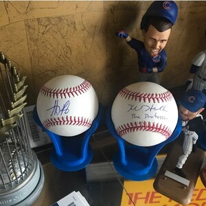 May include: Two white baseballs with blue stitching and black signatures on a blue stand. The baseballs are signed by "Kris Bryant" and "J. Russell The Professor". A silver trophy with gold accents is in the background.