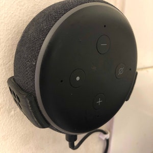 Puede incluir: Un altavoz inteligente circular negro con una cubierta de tela y cuatro botones. El altavoz está montado en una pared con una correa negra y un cable blanco.