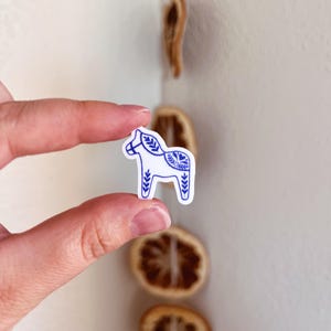 May include: A small white sticker shaped like a Dala horse featuring intricate blue line art patterns on its body, including leaf motifs and a heart design on the flank, presented as a die-cut decal with a clean white border.