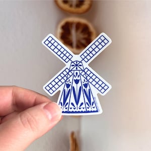 May include: A white sticker featuring a blue and white windmill design. The windmill has four blades and a detailed body with floral patterns. The sticker is held by a hand.
