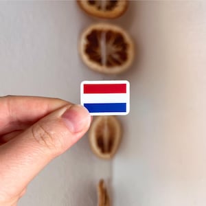 Puede incluir: Una pequeña pegatina rectangular que muestra la bandera tricolor horizontal de los Países Bajos, compuesta por tres franjas iguales de color rojo, blanco y azul. La pegatina tiene esquinas redondeadas y un borde blanco sobre fondo neutro.
