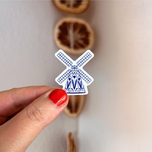Puede incluir: Una pegatina troquelada que presenta una ilustración en azul y blanco de un molino de viento tradicional. El diseño del molino incluye un patrón de cuadrícula en las aspas y una base decorativa adornada con motivos florales de tulipanes.
