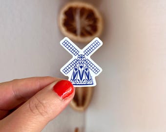 May include: A die-cut sticker featuring a blue and white illustration of a traditional windmill. The windmill design includes a grid pattern on the blades and a decorative base adorned with stylized tulip floral motifs in a deep blue ink.