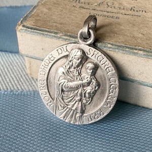 Könnte beinhalten: Silberfarbener Anhänger mit detailliertem Relief einer Frau, die ein Kind hält. Der Anhänger hat eine runde Form mit Text am Rand. Der Hintergrund umfasst eine Vintage-Box mit Text.