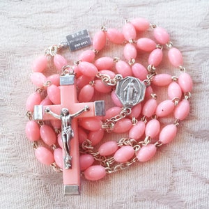 May include: A pink rosary with oval beads and silver-tone metal chain. The rosary features a pink cross with a silver-tone crucifix and a silver-tone medal with a religious figure.