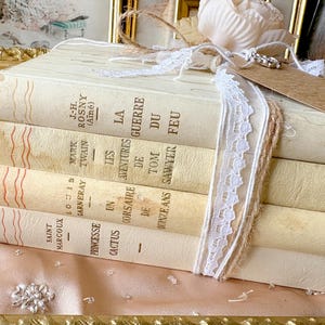 Può includere: Una pila di libri d'epoca con copertine color crema invecchiate, legati con spago e pizzo bianco. I titoli sono in francese. Un piccolo tag e un fiore decorativo sono attaccati, suggerendo un oggetto decorativo o regalo.
