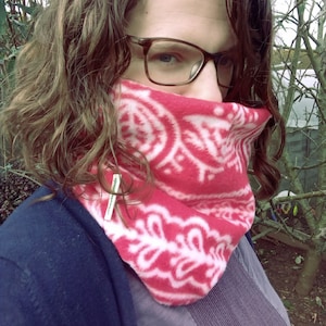 Puede incluir: Un cuello de forro polar con estampado rojo y blanco. El cuello está envuelto alrededor del cuello y cubre la parte inferior de la cara. La persona lleva gafas y un jersey azul. El forro polar tiene un diseño floral blanco.