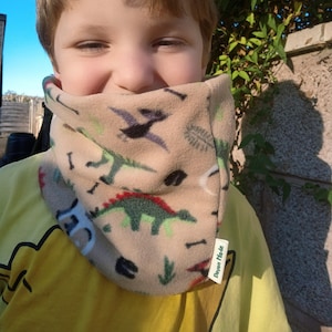 May include: A young boy wearing a tan fleece neck warmer with a dinosaur and leaf print. The neck warmer has a green label that says "Devon Made".