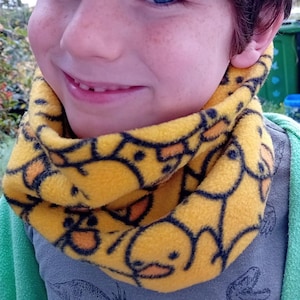 Puede incluir: Un cuello polar amarillo con un patrón repetido de patos de dibujos animados con contornos negros. El cuello se lleva alrededor del cuello de una persona. La persona lleva una camiseta gris y una chaqueta verde.