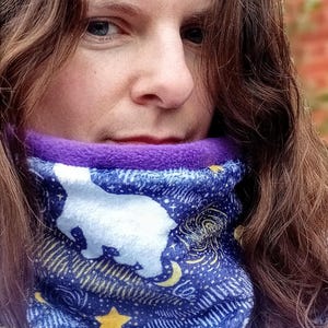 Peut inclure: Une femme aux longs cheveux bruns porte un cache-cou de couleur violet et bleu avec un ours polaire blanc et des étoiles.