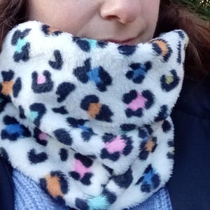 以下が含まれることがあります： 白のフェイクファーネックウォーマーにレオパード柄とカラフルなアクセントが施されています。ネックウォーマーは折り畳んで首に巻かれています。
