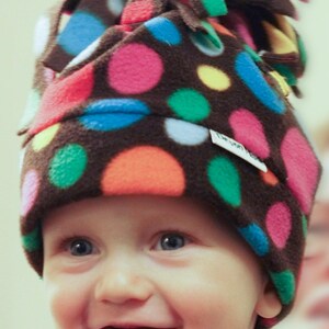 May include: A brown fleece hat with colourful polka dots in pink, orange, blue, green, and yellow. The hat has a unique design with layered fabric at the top. The child wearing the hat is smiling.