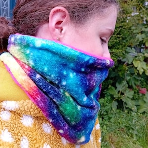 Puede incluir: Un calentador de cuello colorido con estampado de galaxia. El calentador de cuello está hecho de una tela suave y peluda y tiene un forro rosa.