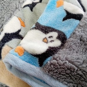 Puede incluir: Una manta suave y mullida con un patrón de pingüinos azul y blanco. La manta está doblada en tres partes, revelando un forro de sherpa gris.