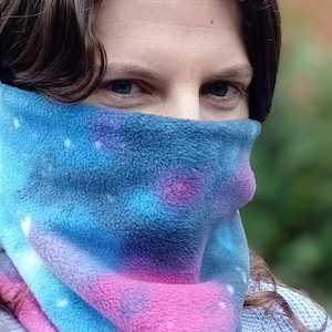 Puede incluir: Una mujer lleva un cuello de lana azul, rosa y blanco con estampado tie-dye. El cuello de lana es suave y acogedor, perfecto para mantenerse caliente en invierno.