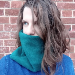 Puede incluir: Un calentador de cuello de forro polar verde azulado se usa alrededor del cuello, cubriendo la parte inferior de la cara. La persona lleva una parte superior azul. El fondo es una pared de ladrillos rojos. La persona tiene el pelo castaño.