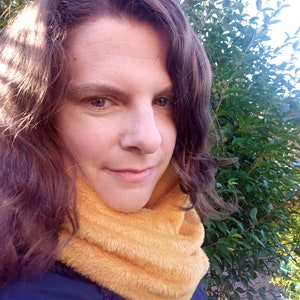 Puede incluir: Una mujer con el pelo largo y castaño lleva una chaqueta azul y un cuello de lana amarillo. Está de pie frente a un arbusto verde y frondoso.